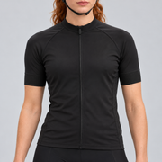 Female model wearing the plain black short-sleeve GG BASICS cycling jersey with black collar, cuffs and side panels, shown with black cycling shorts against a light grey studio background.