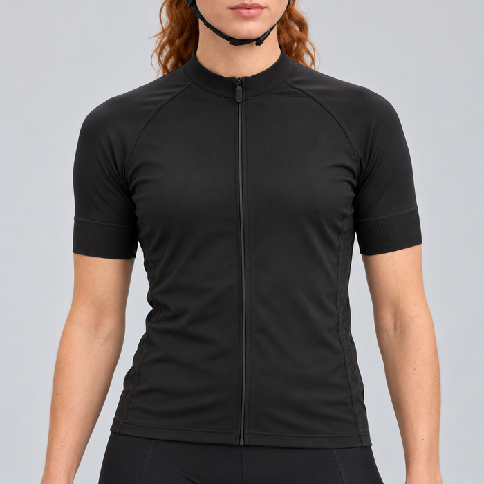 Female model wearing the plain black short-sleeve GG BASICS cycling jersey with black collar, cuffs and side panels, shown with black cycling shorts against a light grey studio background.
