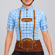 Female model wearing the pale blue and white checked short-sleeve Blue Lederhosen cycling jersey with printed brown braces, floral folk motifs and edelweiss sleeve details, shown with black cycling shorts against a light grey studio background.