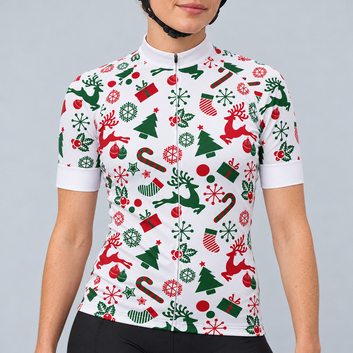 Female model wearing the white short-sleeve Christmas cycling jersey with red and green reindeer, trees, snowflakes, stockings, presents and candy canes, shown with black cycling shorts against a light grey studio background.