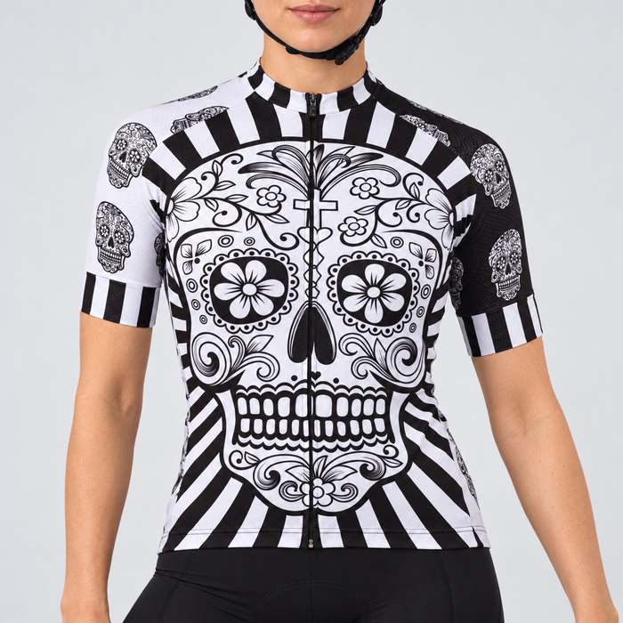 Female model wearing the black-and-white short-sleeve Day Of The Dead Skull cycling jersey with a large calavera skull and radiating stripe design, shown with black cycling shorts against a light grey studio background.