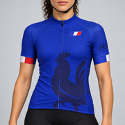 Female model wearing the royal-blue short-sleeve France cycling jersey with a tonal rooster graphic, French tricolour chest patch and flag-trimmed cuffs, shown with black cycling shorts against a light grey studio background.