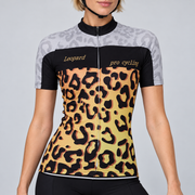 Female model wearing the short-sleeve Leopard Print cycling jersey with pale grey shoulders, black chest panel, gold and black leopard-print body, and black cycling shorts against a light grey studio background.