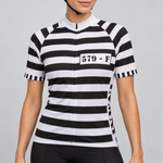 Female model wearing the black-and-white striped short-sleeve Prison Inmate cycling jersey with a 579 - F chest number panel, shown with black cycling shorts against a light grey studio background.