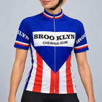 Female model wearing the royal blue, red and white Retro Brooklyn short-sleeve cycling jersey with BROO KLYN CHEWING GUM chest lettering, shown with black cycling shorts against a light grey studio background.