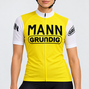 Female model wearing the yellow and white short-sleeve Dr. Mann Grundig retro cycling jersey with black MANN GRUNDIG lettering and black cycling shorts against a light neutral studio background.