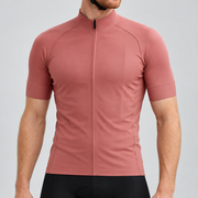 Male model wearing the plain rose-gold short-sleeve GG BASICS cycling jersey with matching collar and cuffs and mesh side panels, shown with black cycling shorts against a light grey studio background.